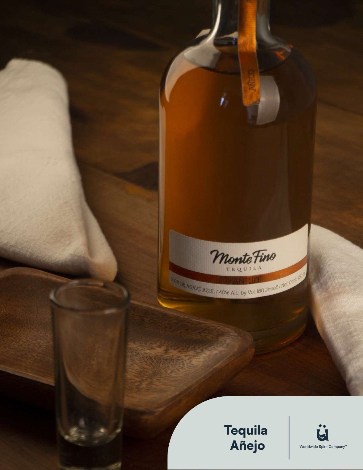 Bottle of Monte Fino Añejo tequila on a wooden table with a shot glass and white cloth napkins.