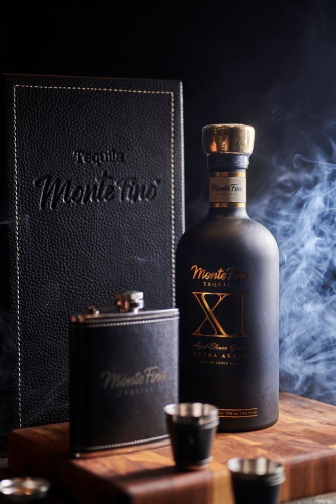 Dark liquor bottle, flask, and shot glasses on a wooden board. Black and gold design. Smoke effect.