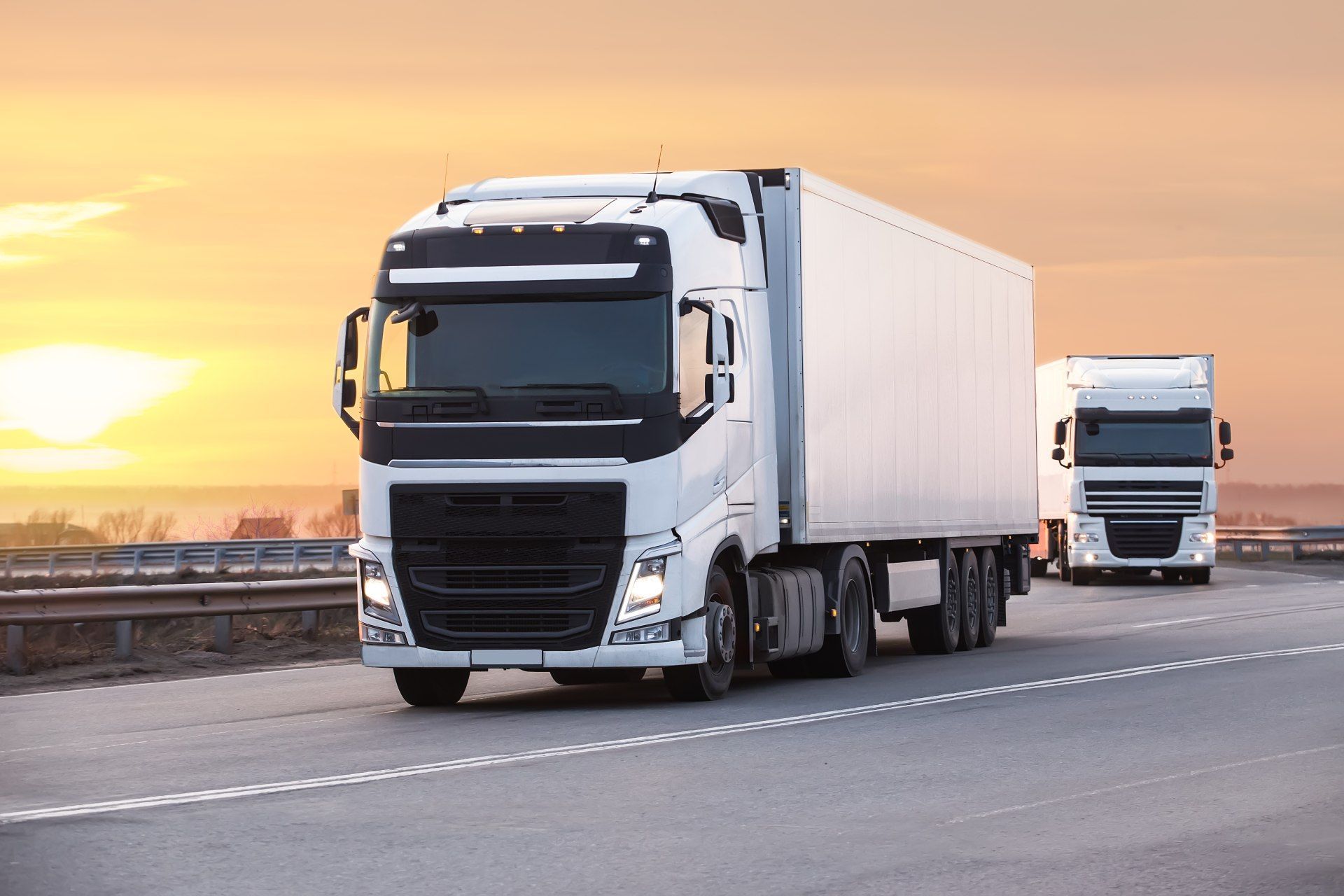 Two white semi-trucks driving on a highway against a sunset background.