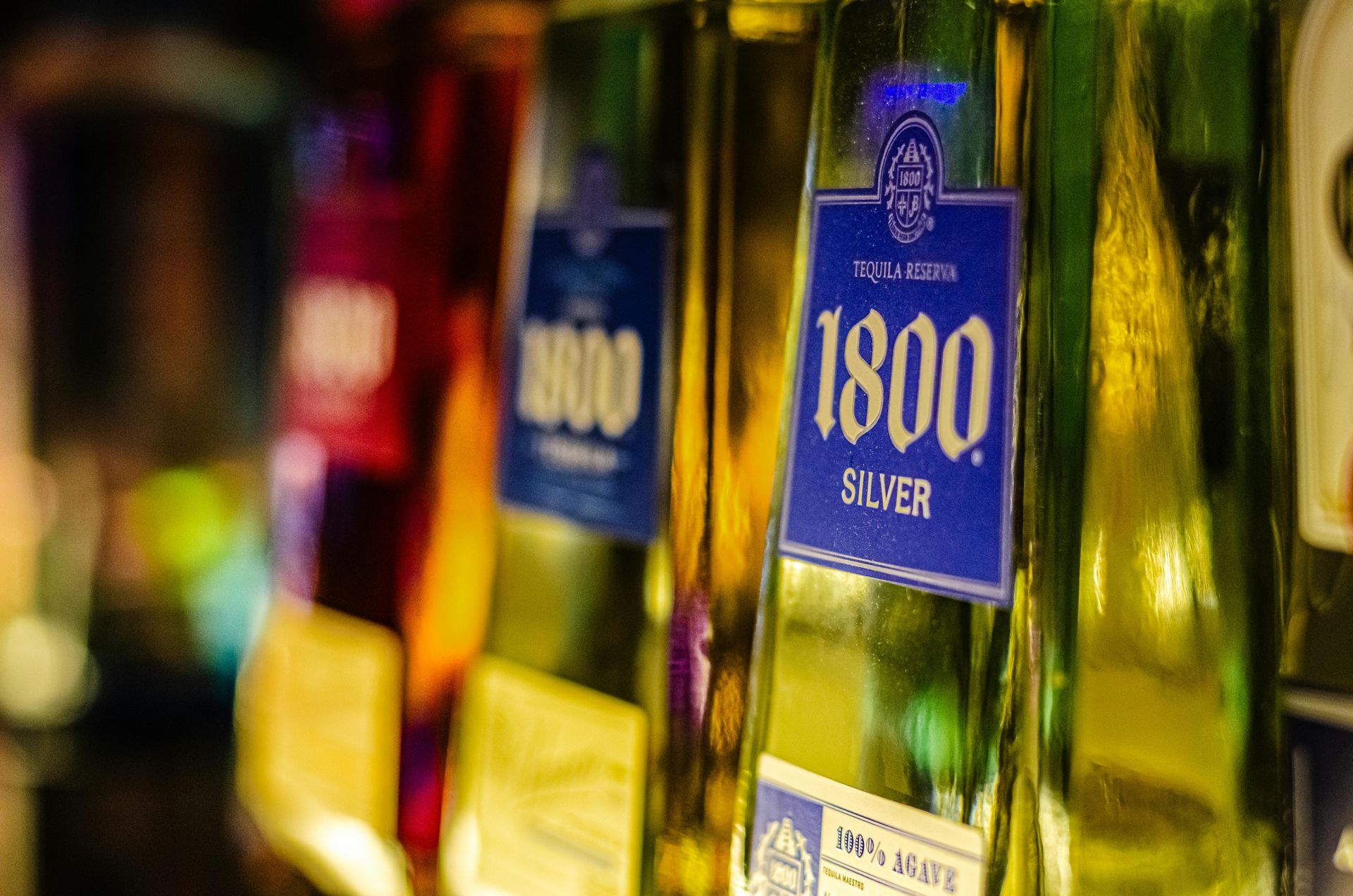 Tequila bottles in a row, a silver 1800 label is visible, with colorful bottles beside it, inside a bar.