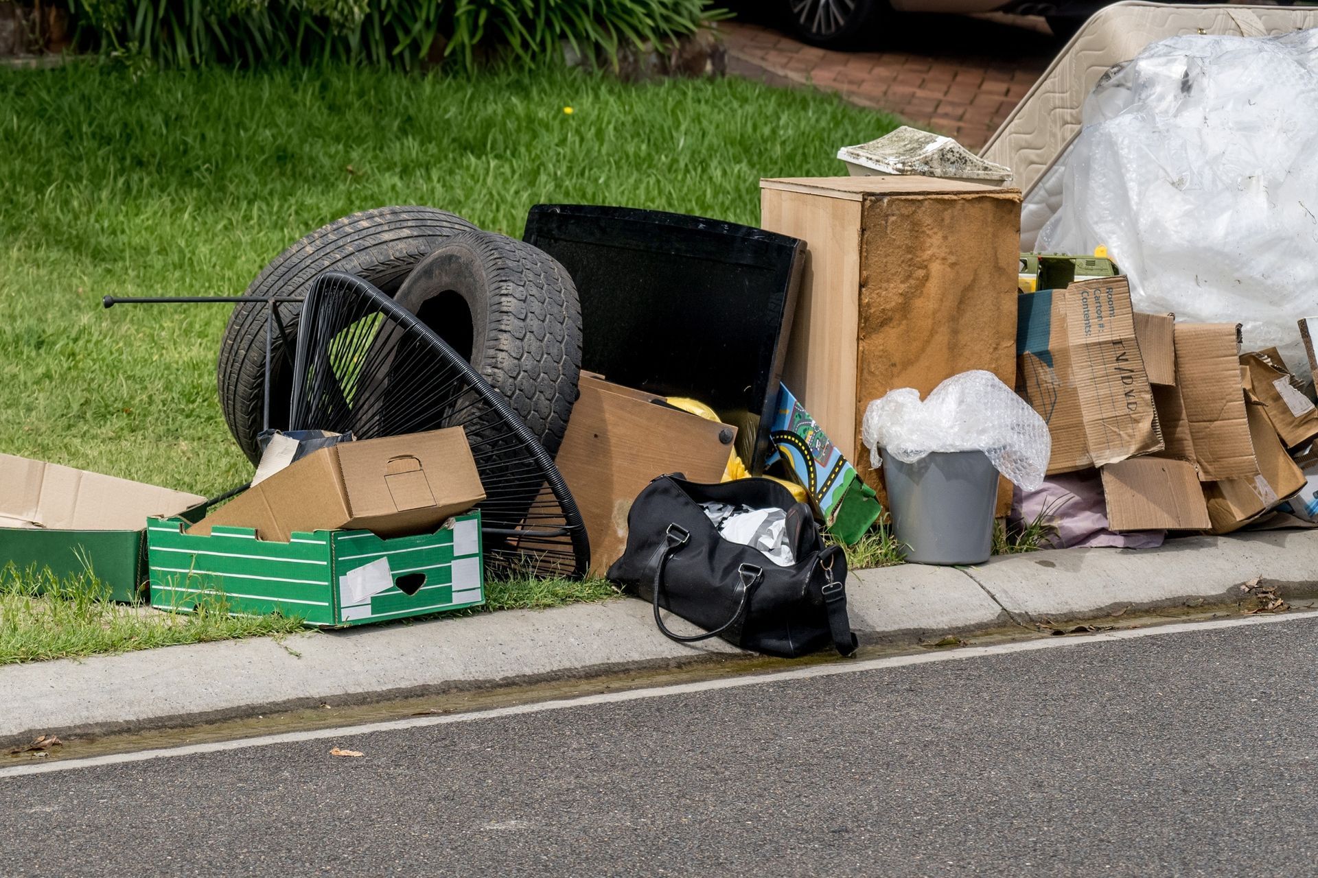 Pile of discarded trash, including tires, cardboard, and a bag, on a curb next to grass.