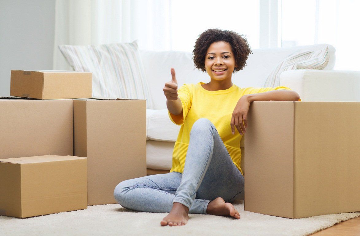 A woman is sitting on the floor surrounded by cardboard boxes and giving a thumbs up.