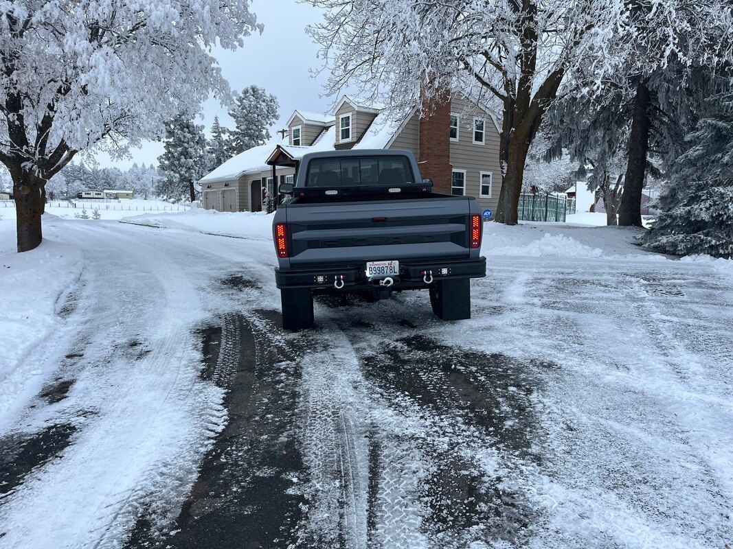 A truck is driving down a snowy road in front of a house.