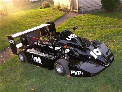 A black race car is parked in the grass in front of a house.