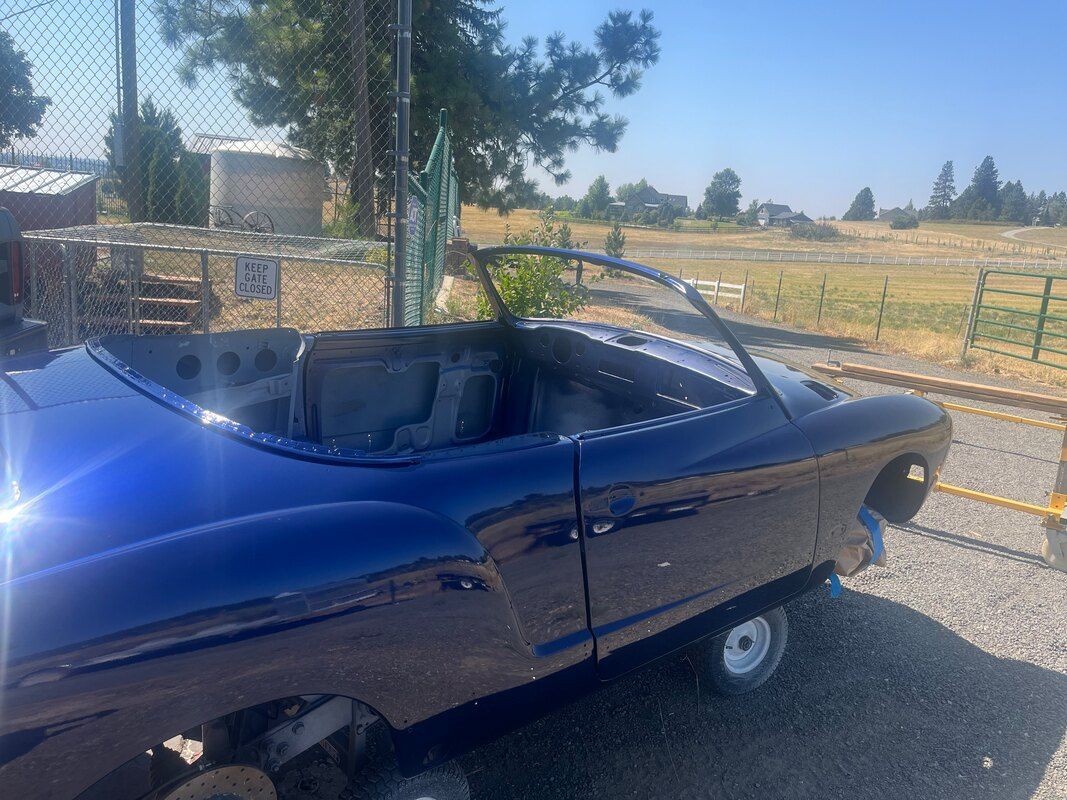 A blue convertible car is parked in a gravel lot next to a fence.