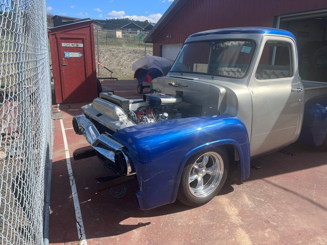 A silver and blue truck with the hood up is parked in a driveway.