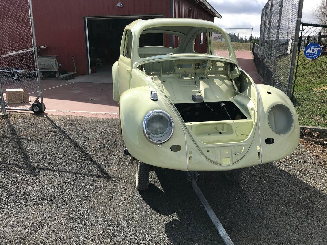 A yellow car is parked in a gravel lot in front of a red barn.