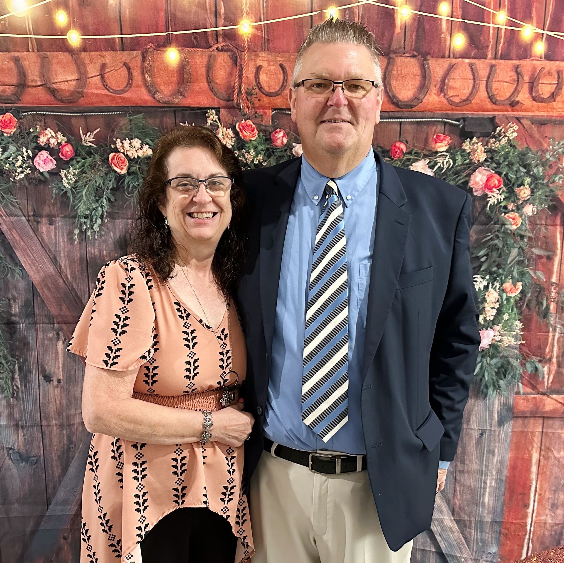 Smiling couple posing in front of a floral and wood backdrop, man in suit and tie, woman in patterned dress