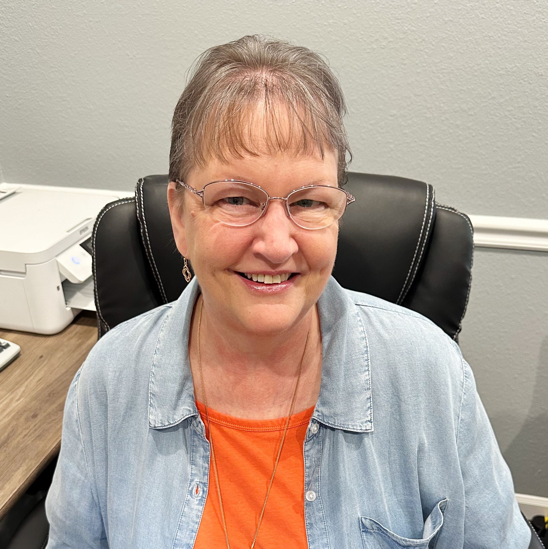 Smiling woman in glasses seated in an office chair, wearing a light denim shirt and orange top.