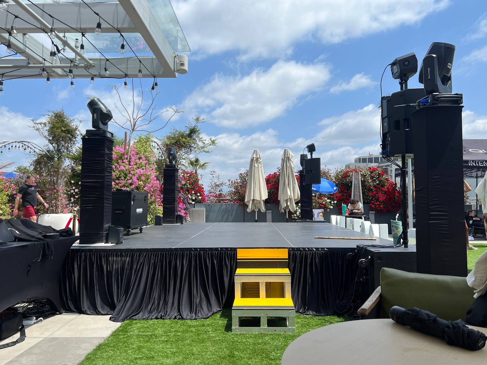 Outdoor stage setup with yellow steps, black drapes, speakers, and floral decorations under a blue sky