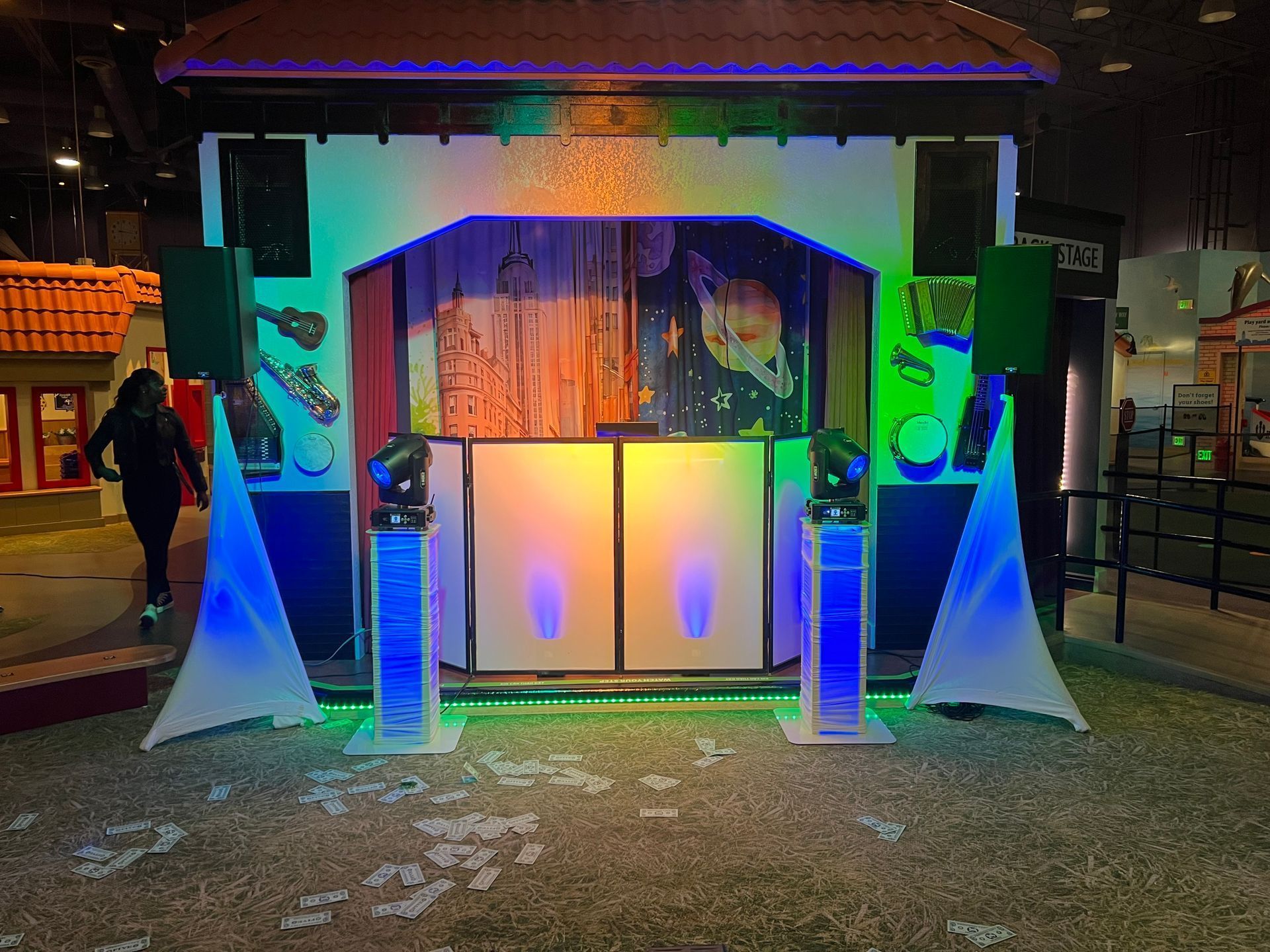 Indoor art exhibit with a small house-like display, colorful lights, and wooden benches on artificial grass