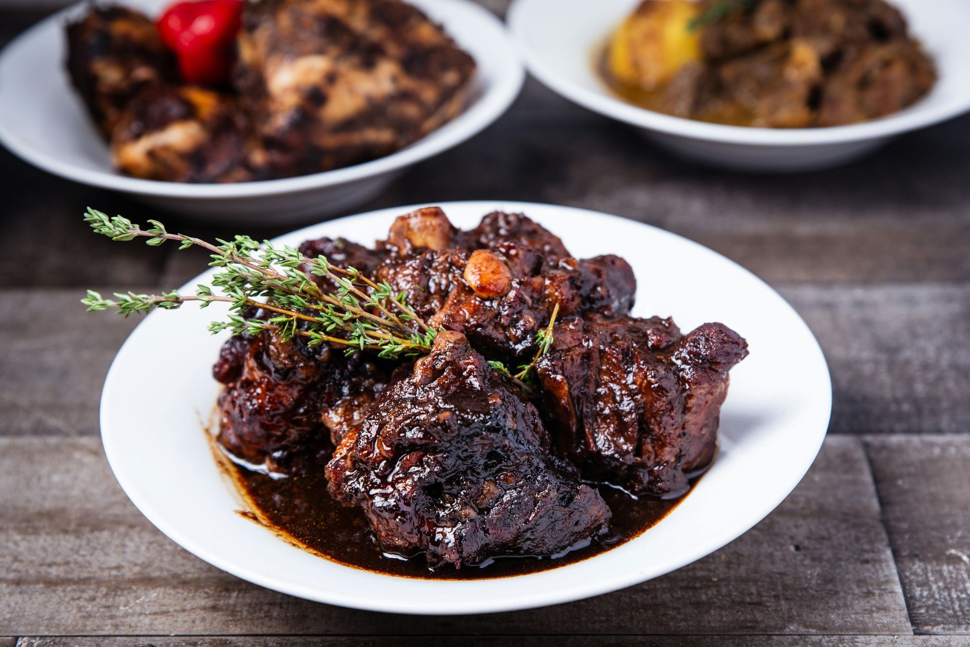Oxtail stew in a white bowl, garnished with thyme; other dishes in background.
