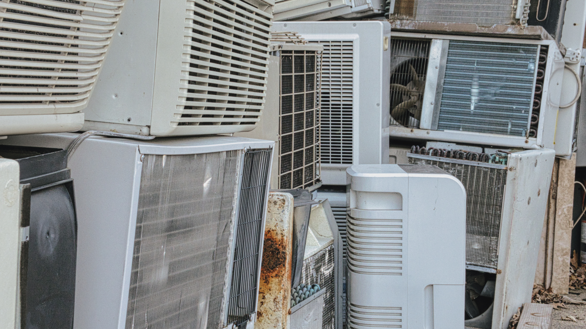 Pile of old, weathered air conditioning units, likely discarded in an outdoor setting.