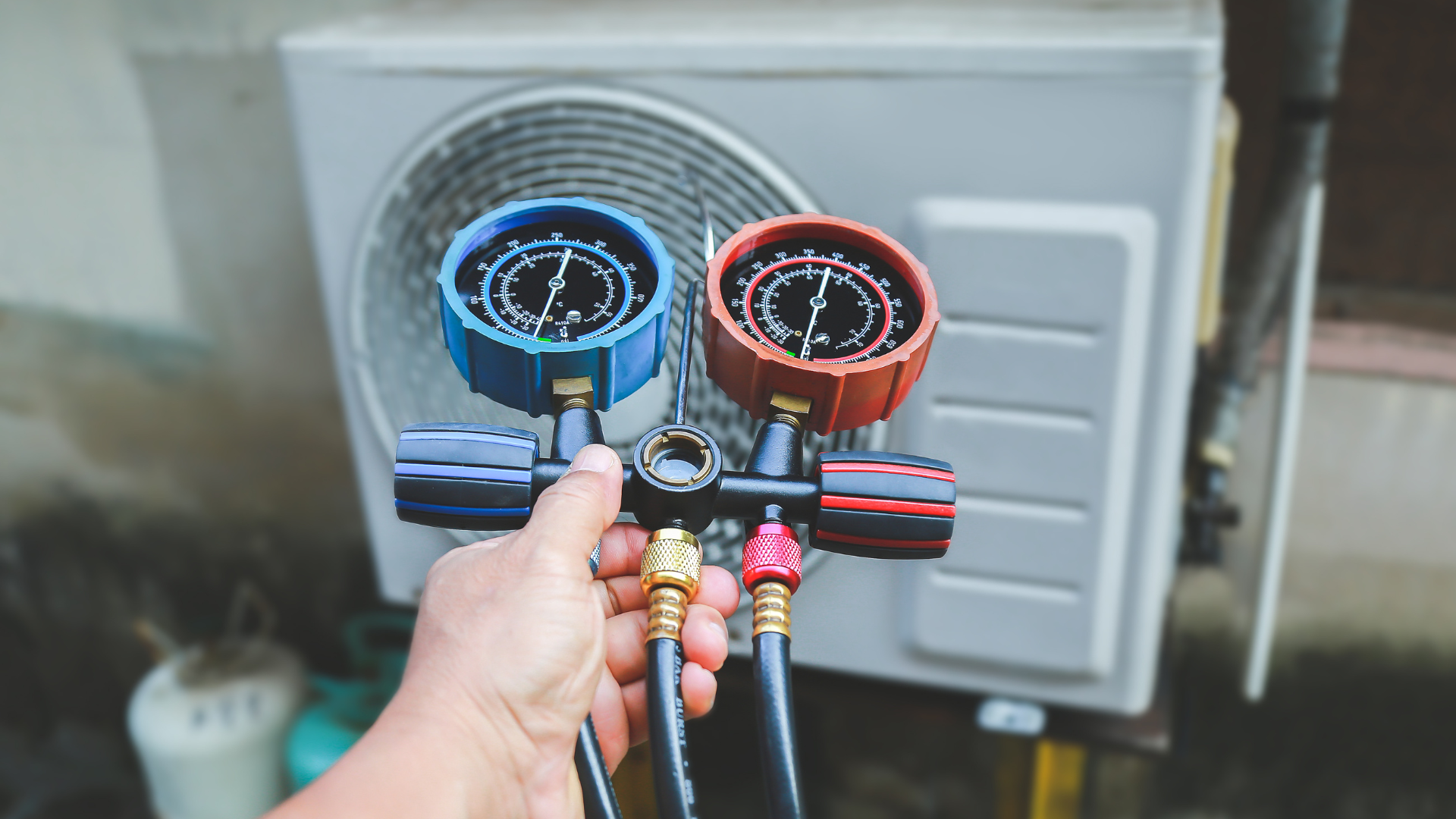 Hand holding gauges connected to an air conditioner unit, presumably for servicing.