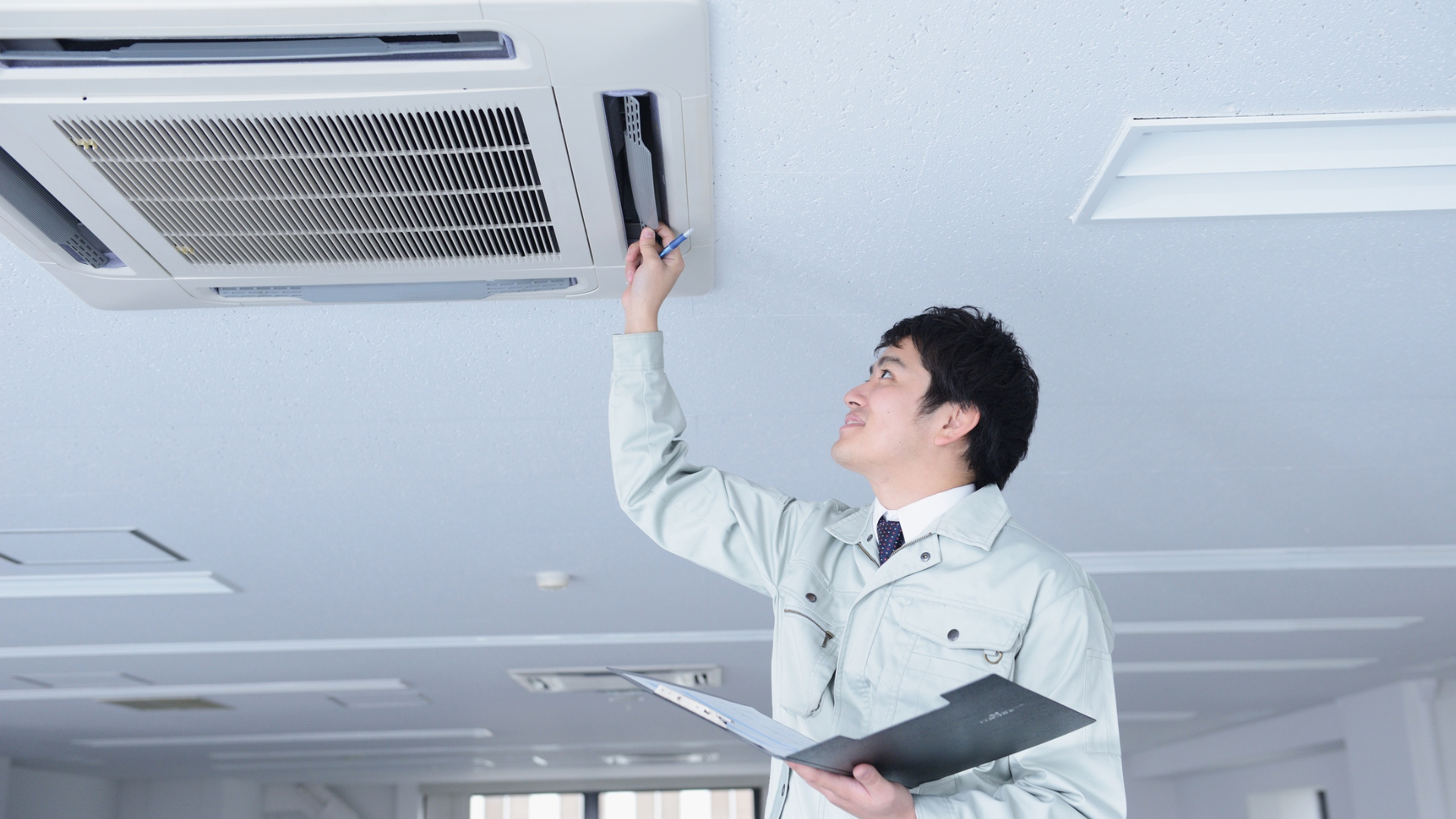 Technician inspecting ceiling-mounted air conditioner in office; holding clipboard.