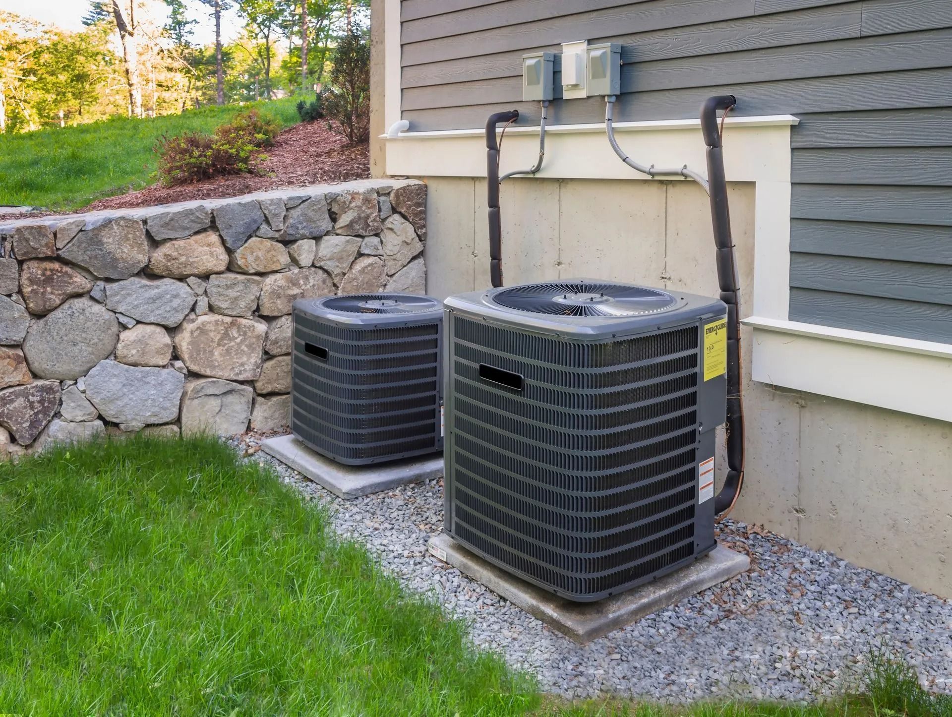 Two outdoor air conditioning units next to a house wall and a stone retaining wall.