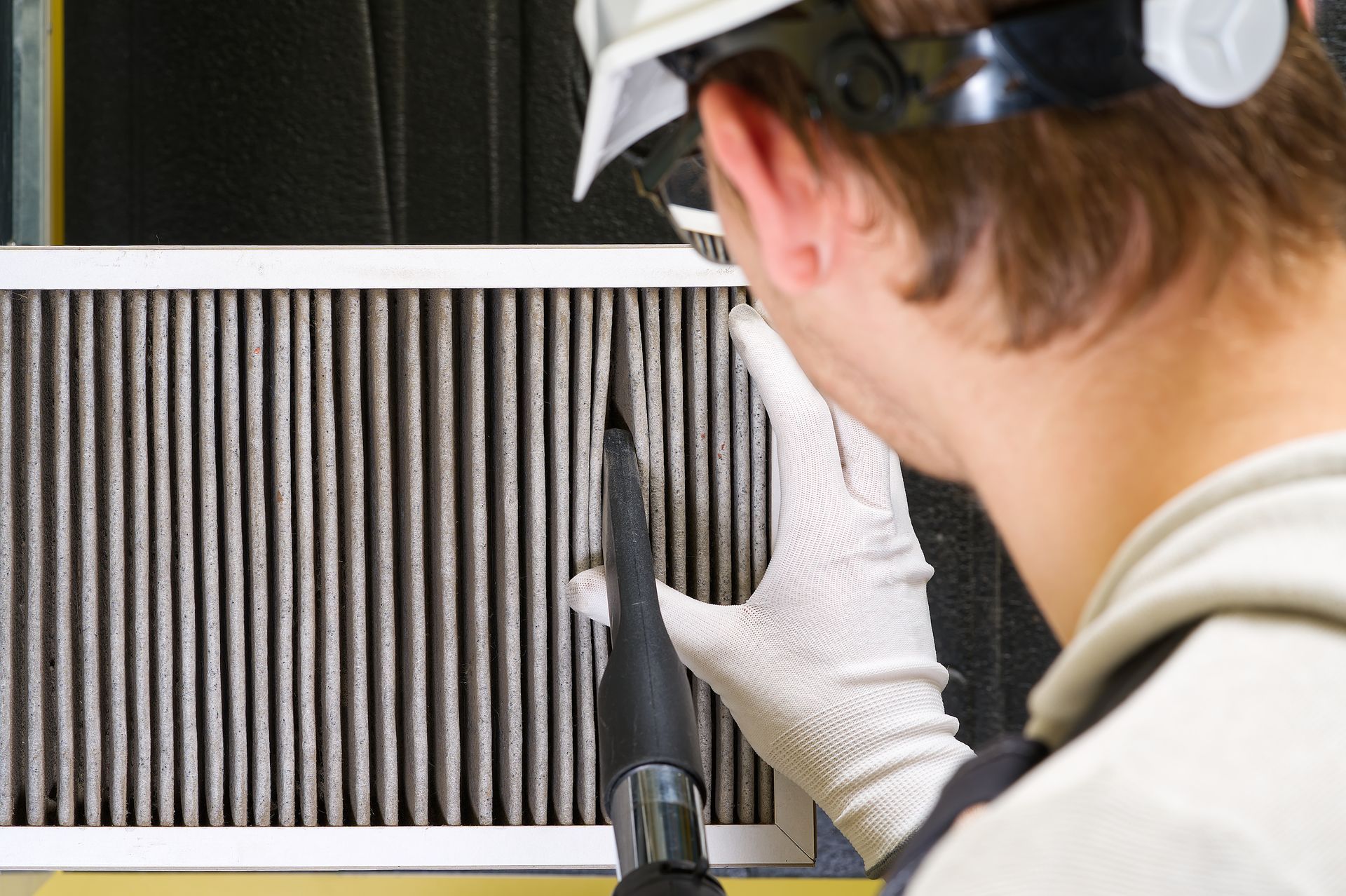 Technician cleaning a dusty air filter with a vacuum, wearing safety glasses and gloves.