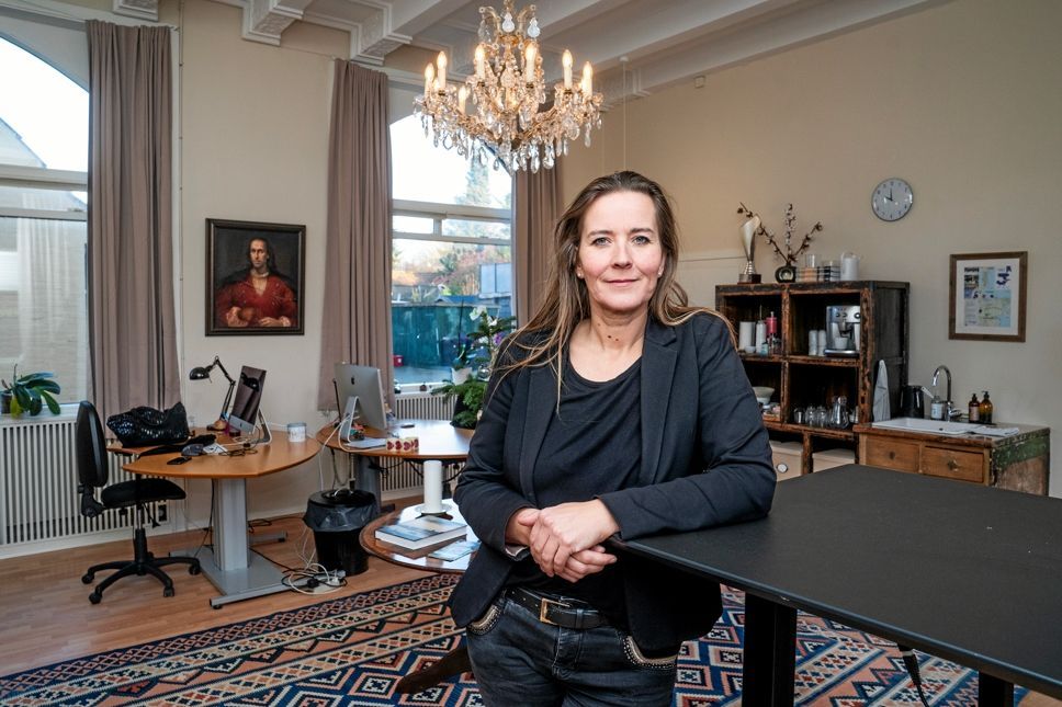 Kvinde i blazer lænet på et bord, på et kontor med en lysekrone, portræt og skrivebord.