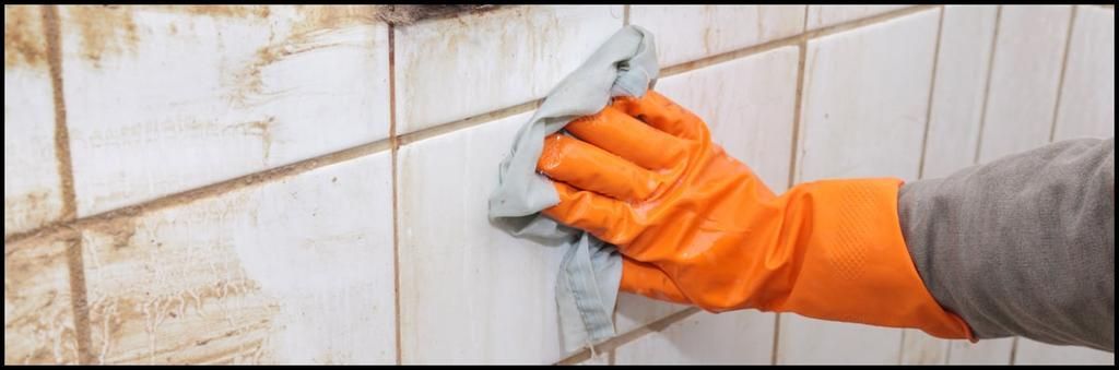 A hand in an orange rubber glove uses a gray cloth to wipe dirt from white bathroom wall tiles.