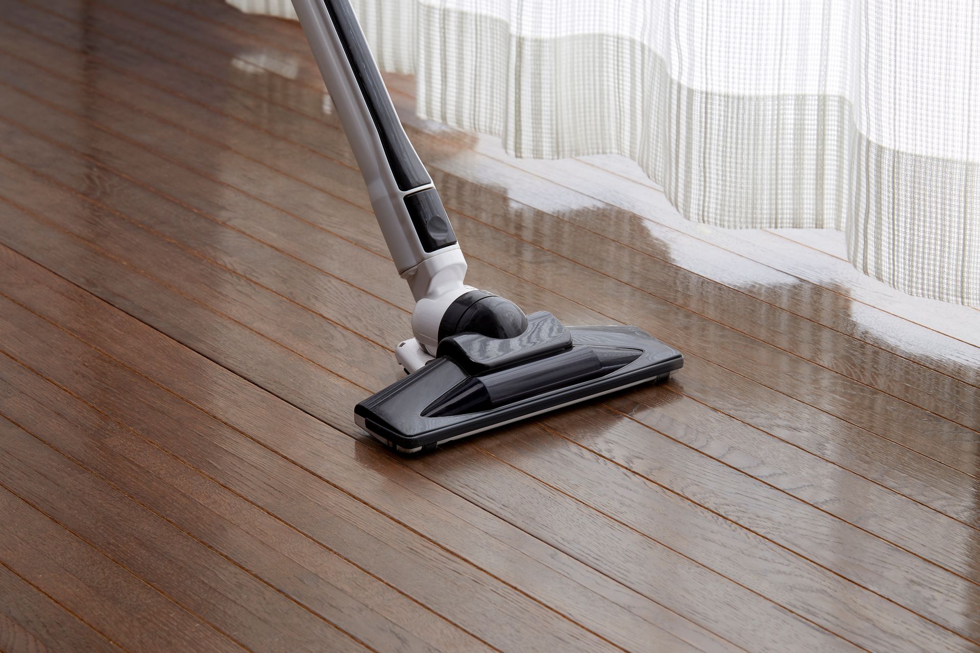 A vacuum cleaner head cleans a polished wood floor near a white curtain.
