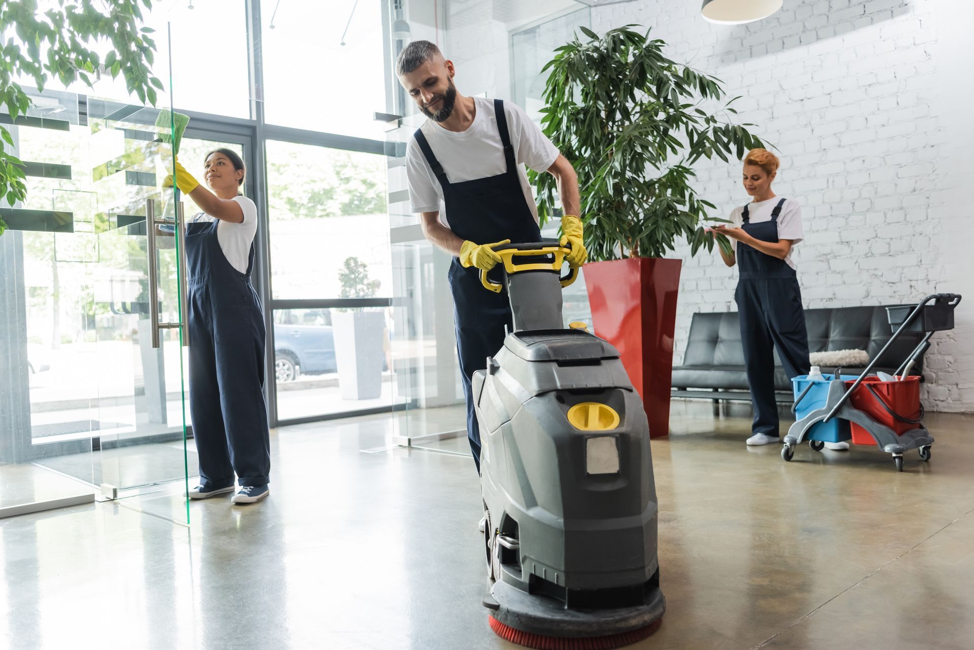 Three professional cleaners in uniforms clean a modern office building, using a floor scrubber and washing a glass door.