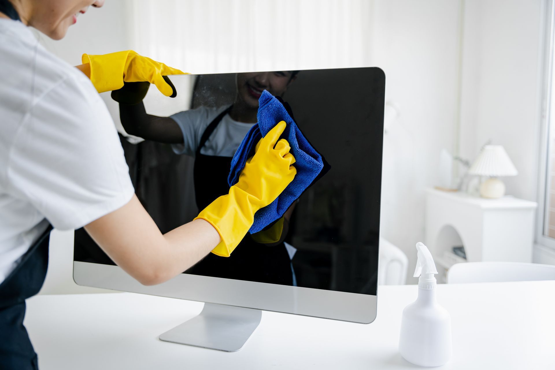 A person wearing yellow cleaning gloves wipes a computer monitor screen.