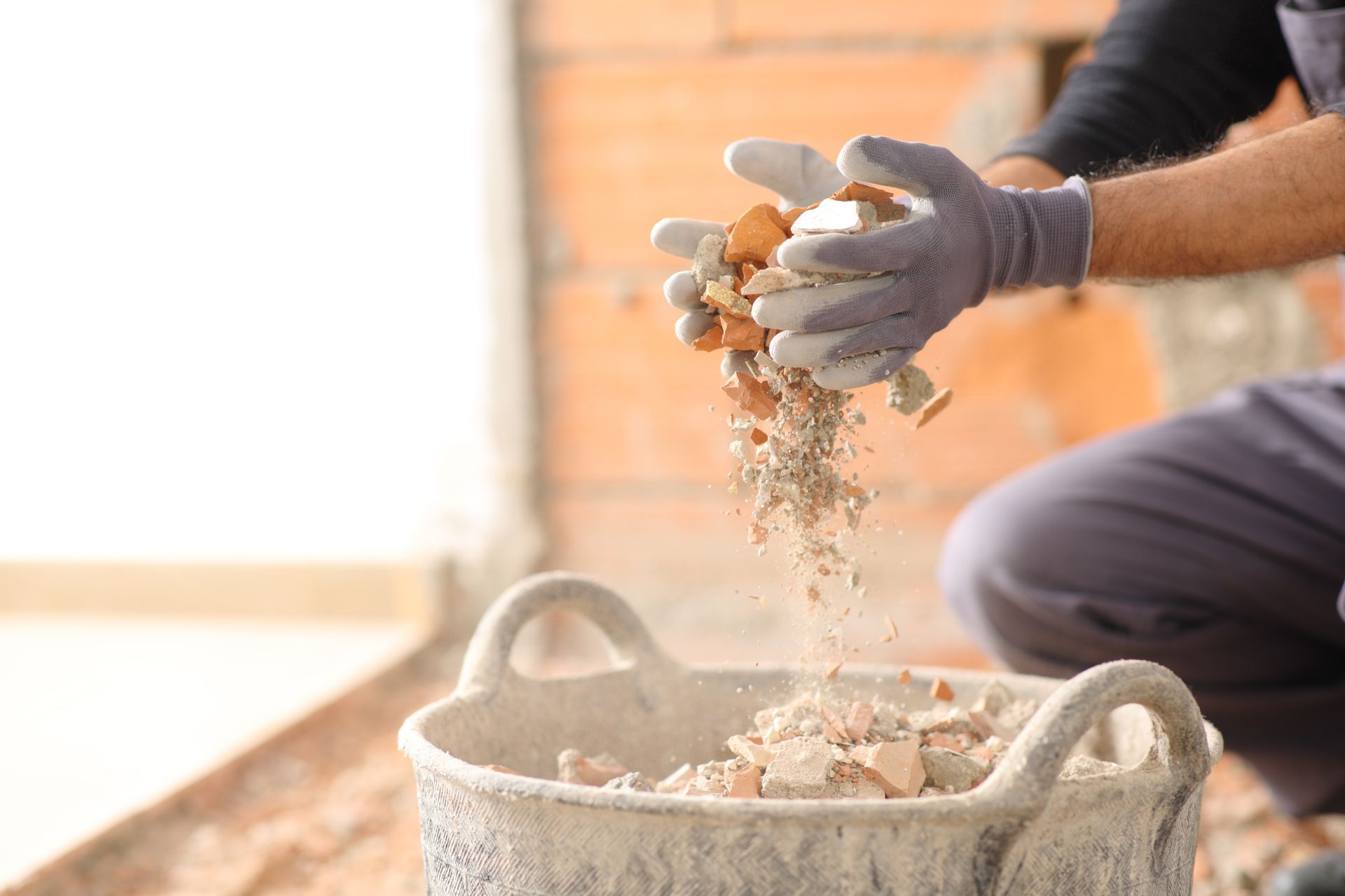 A person wearing work gloves pours construction debris into a plastic bucket on a job site.