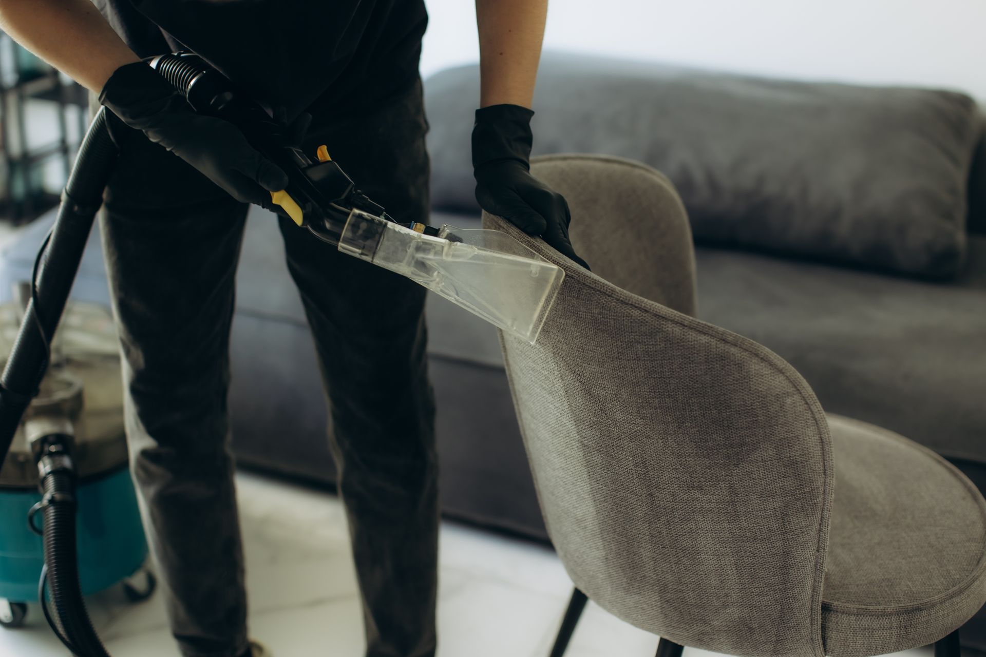 A person in black clothing and gloves uses an upholstery cleaning vacuum.