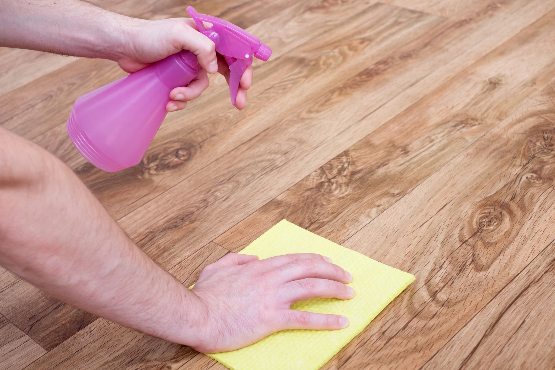 Hands holding a pink spray bottle and wiping a wood floor with a yellow cloth.