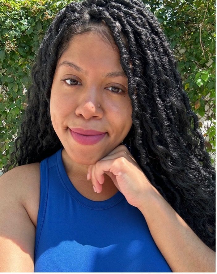 Multiracial woman with long curly hair, slightly smiling, resting her chin on her hands, wearing a royal blue sleeveless top.