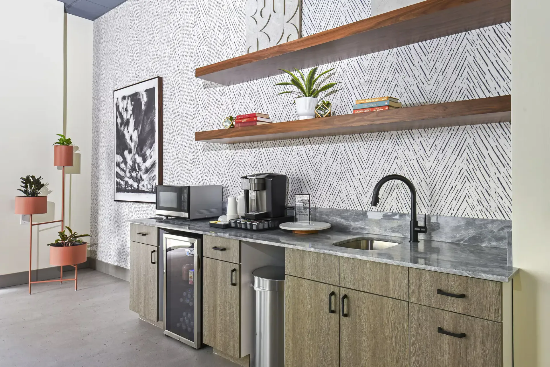 Modern kitchenette with marble countertop, sink, coffee machine, microwave, and wooden shelves.