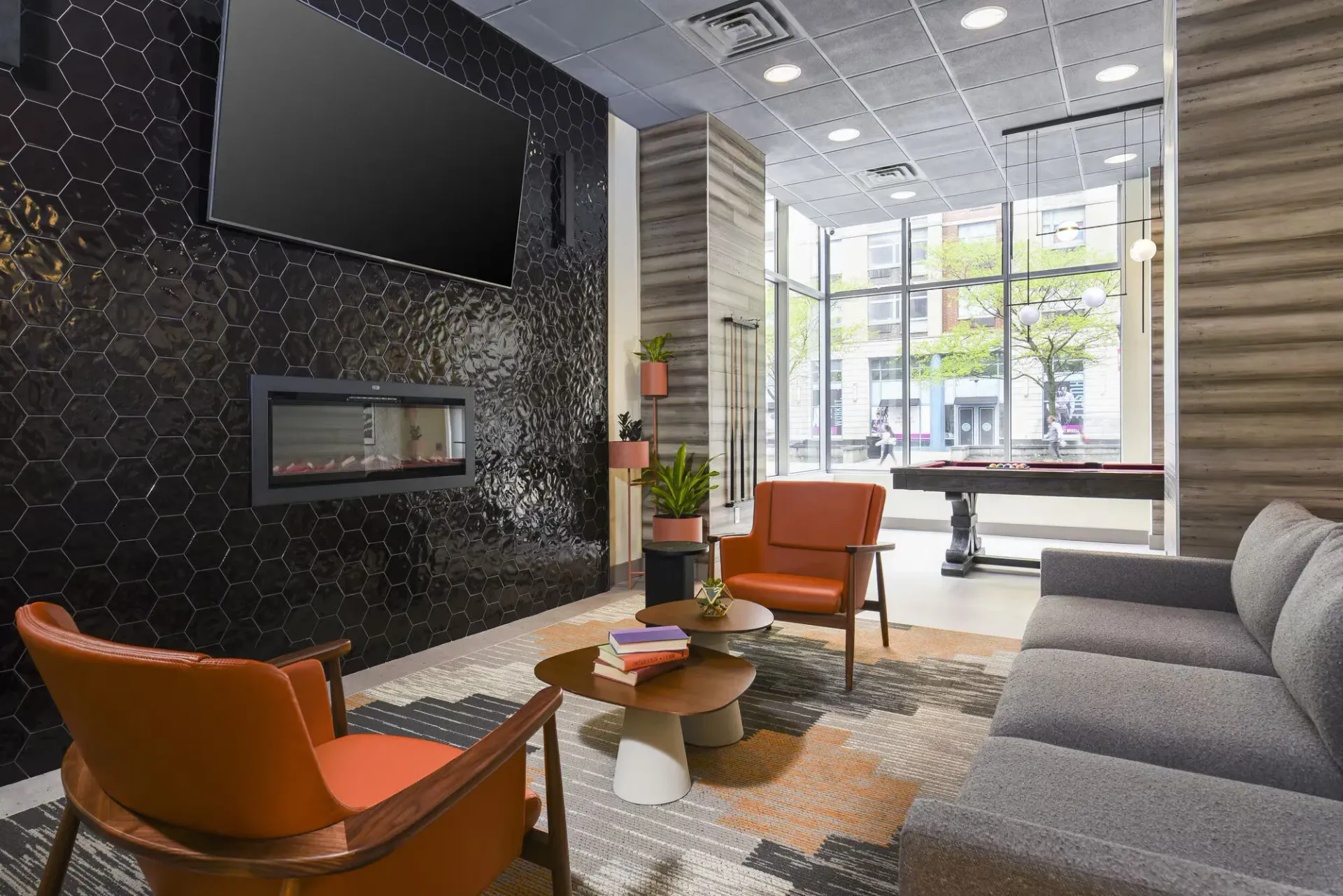Indoor multifamily community lounge with a black hex-tile wall, fireplace, orange chairs, and large window fronts.