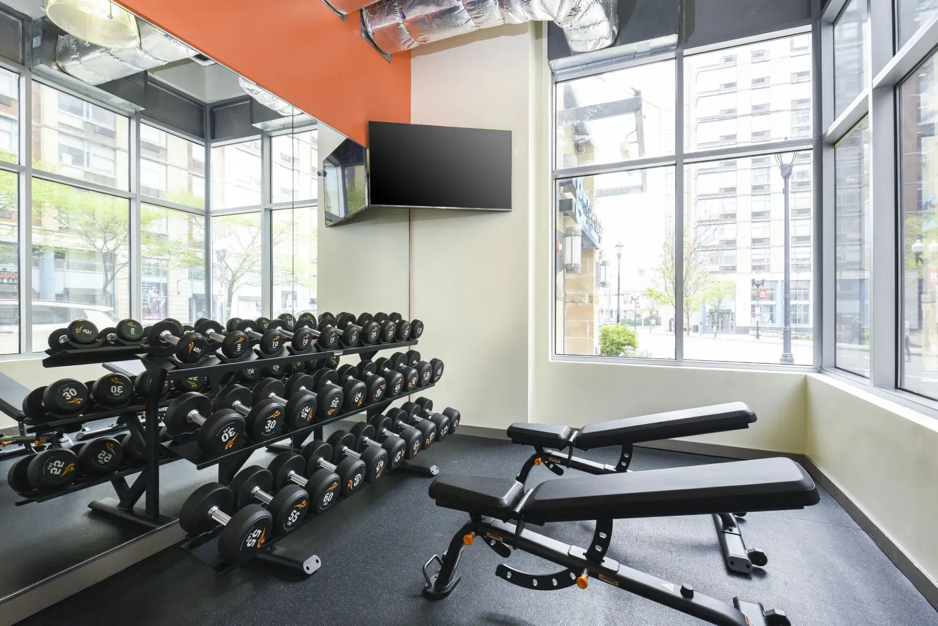 Well-lit gym with dumbbells on racks and a bench near large windows.