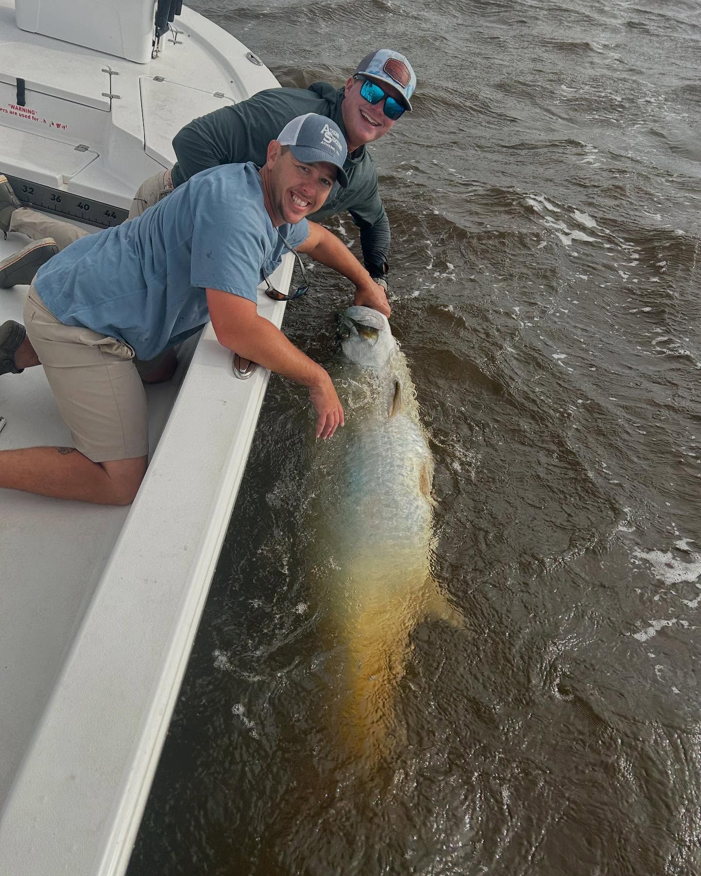 Two men are kneeling on a boat holding a large fish in the water.