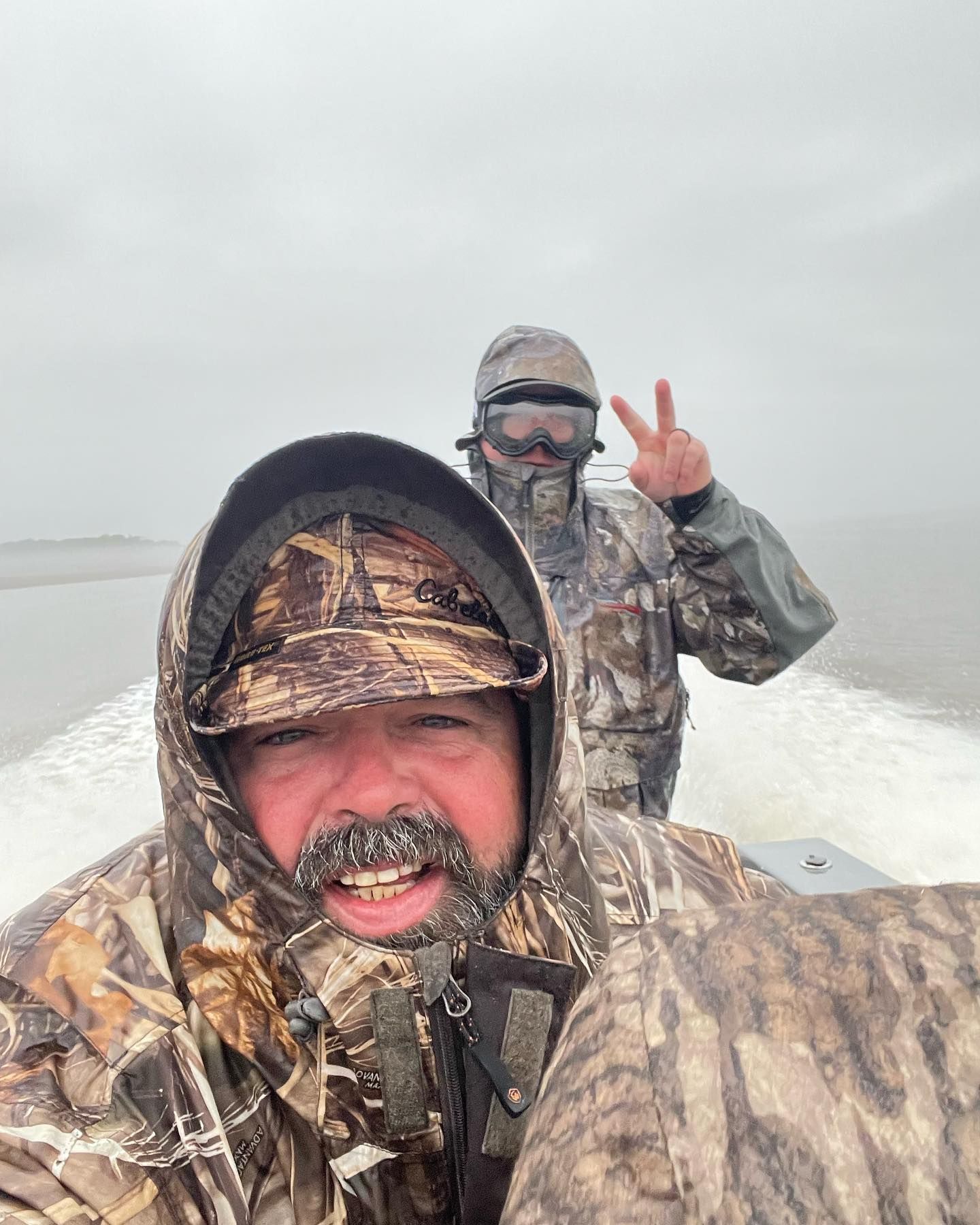 Two men in camo jackets are riding a boat in the water.
