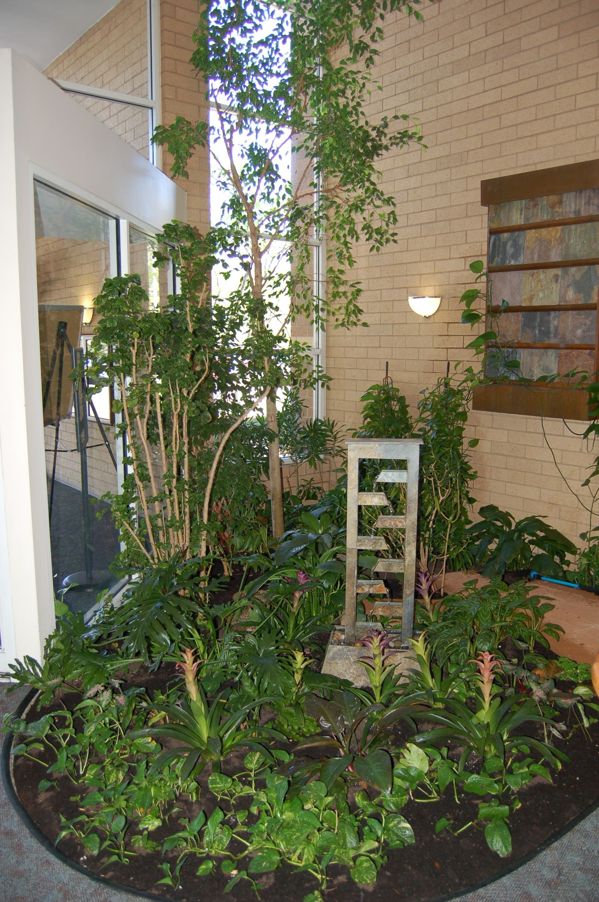 Indoor garden with various plants, a small waterfall, and a brick wall backdrop.