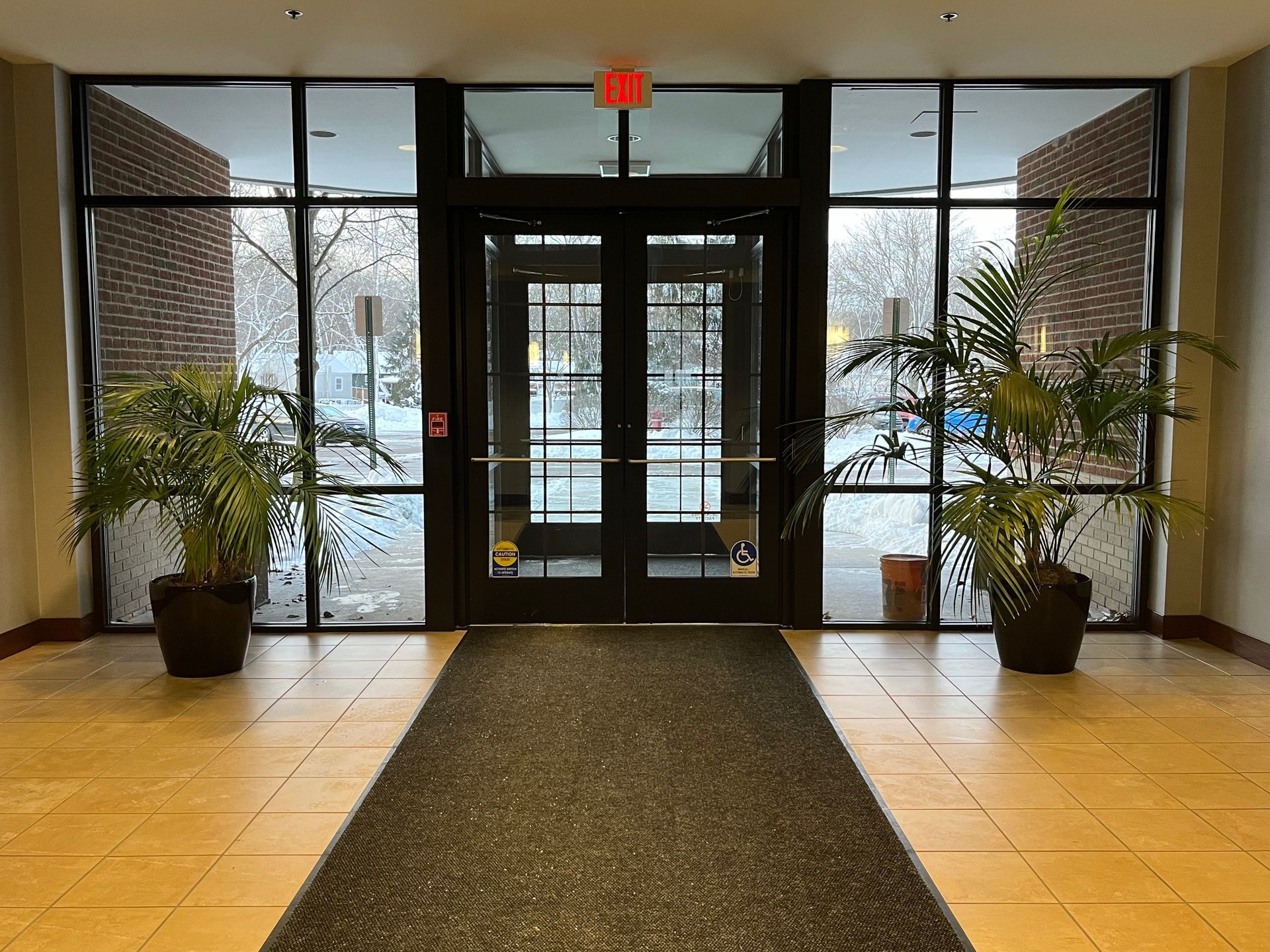Entrance with double doors, windows, potted plants, and a dark carpet. Exterior visible through glass.