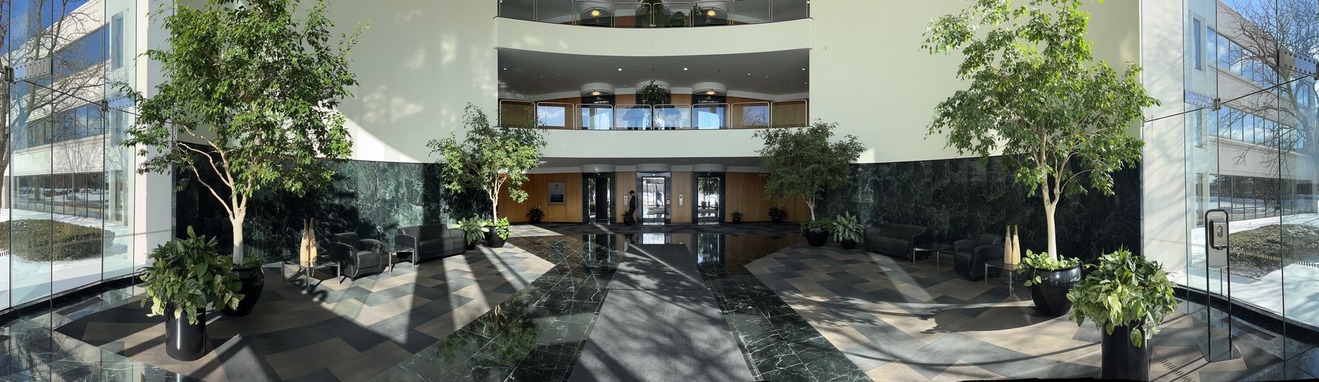 A wide shot of an indoor atrium, featuring trees, plants, and large windows. The interior has a marble floor.