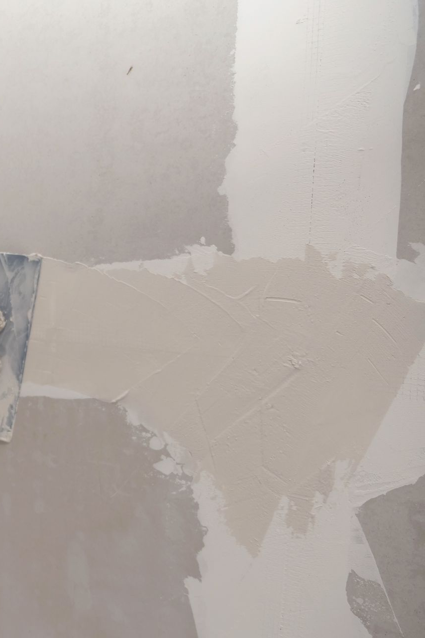 Person using a putty knife to apply drywall mud to a wall, wearing gloves, inside.
