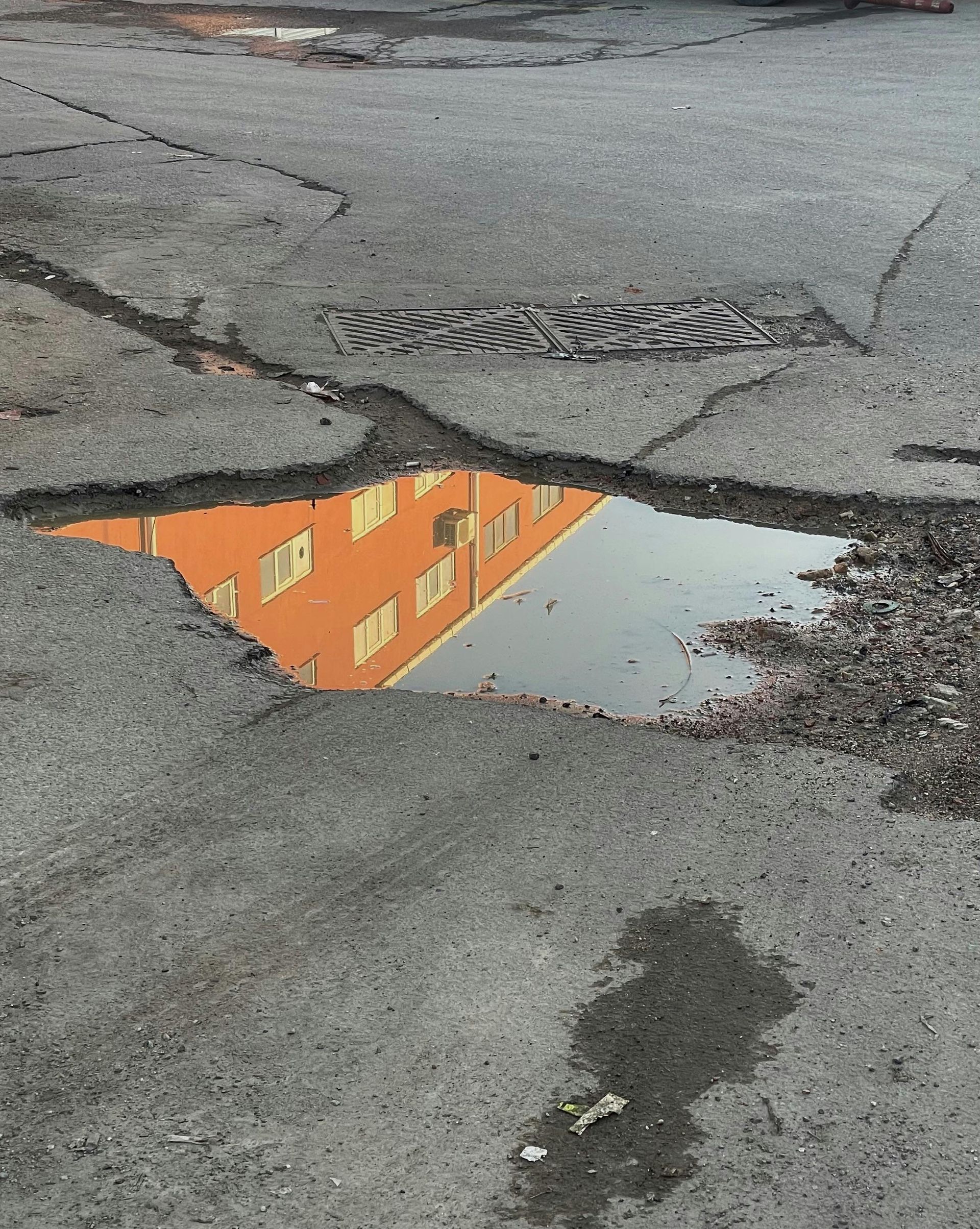 A pothole in gray asphalt reflects an orange building in a pool of standing water.