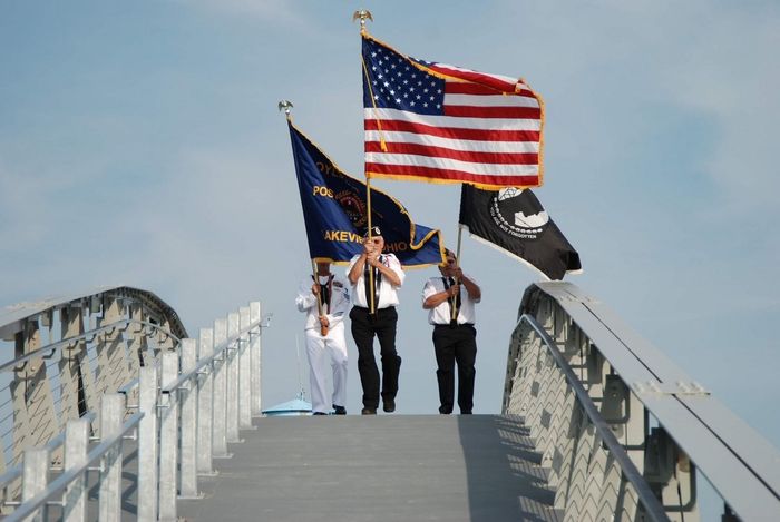 Three people walk across a bridge carrying the American flag, a blue organizational flag, and a black POW/MIA flag.