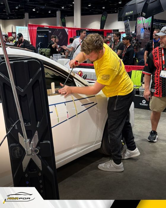 A young man in a yellow shirt works on a car door.  He's at an event with other people in the background.