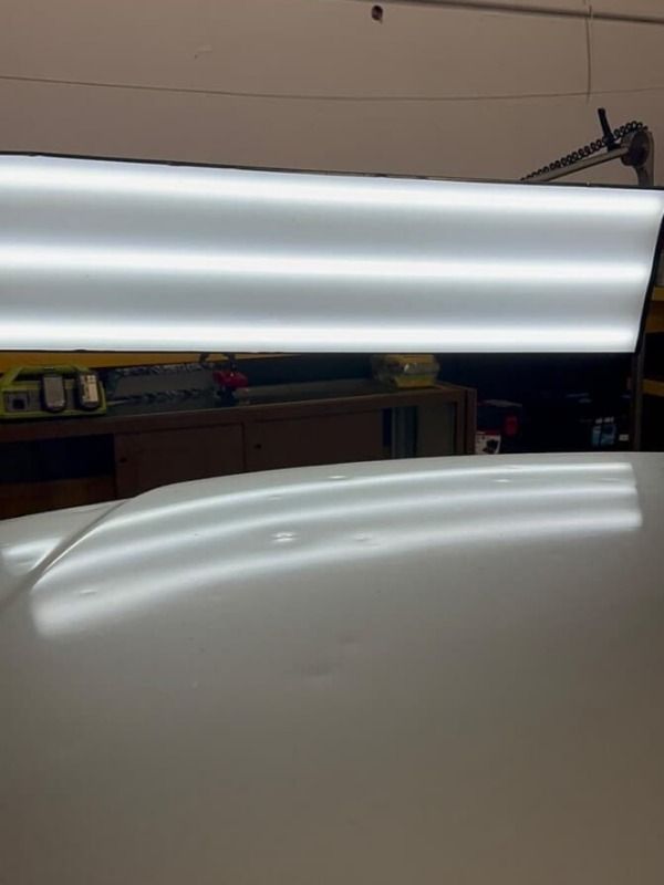 White car roof with dents, illuminated by a bright overhead light panel in a repair shop.