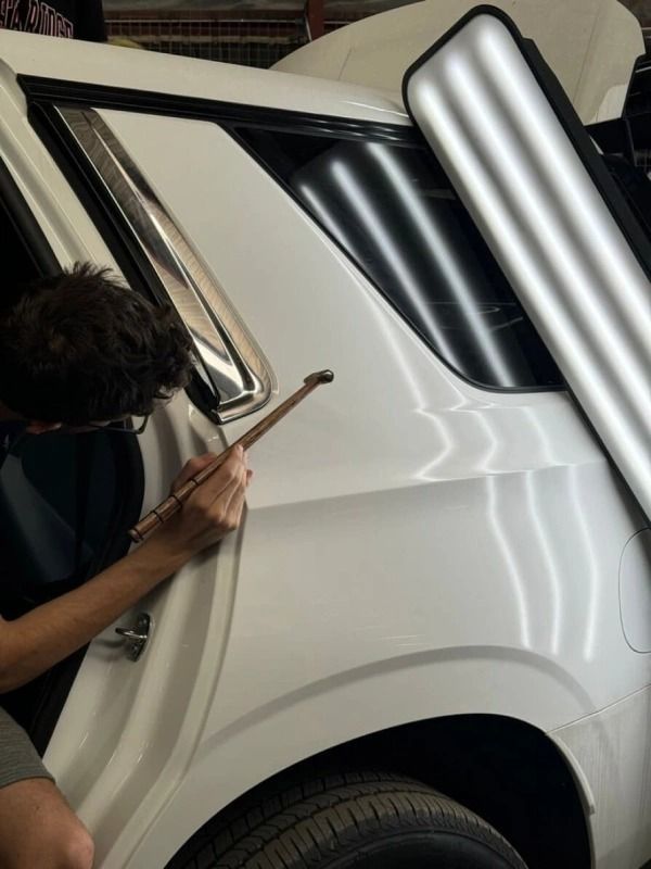 Person using tools to repair a dent on a white car; outdoor setting.