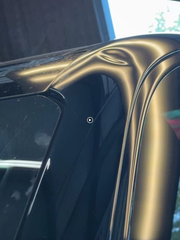 Close-up of a dented, black car roof near a window, reflecting light and revealing the damage.