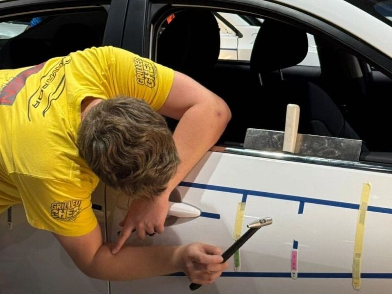 Man hammering car dent, using tools. White car, blue tape markers, yellow shirt.
