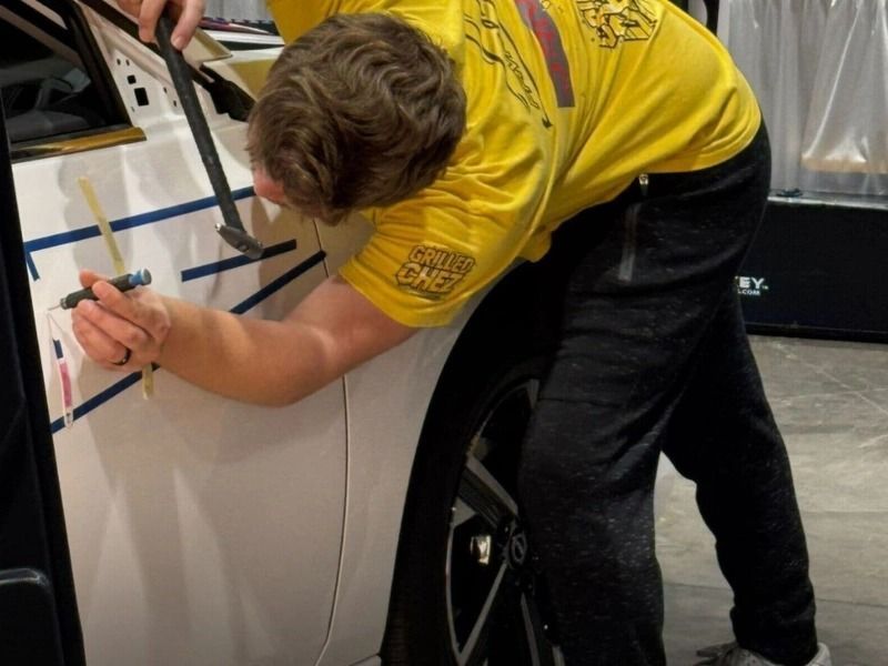 Person in yellow shirt marking lines on a white car door with a hammer.