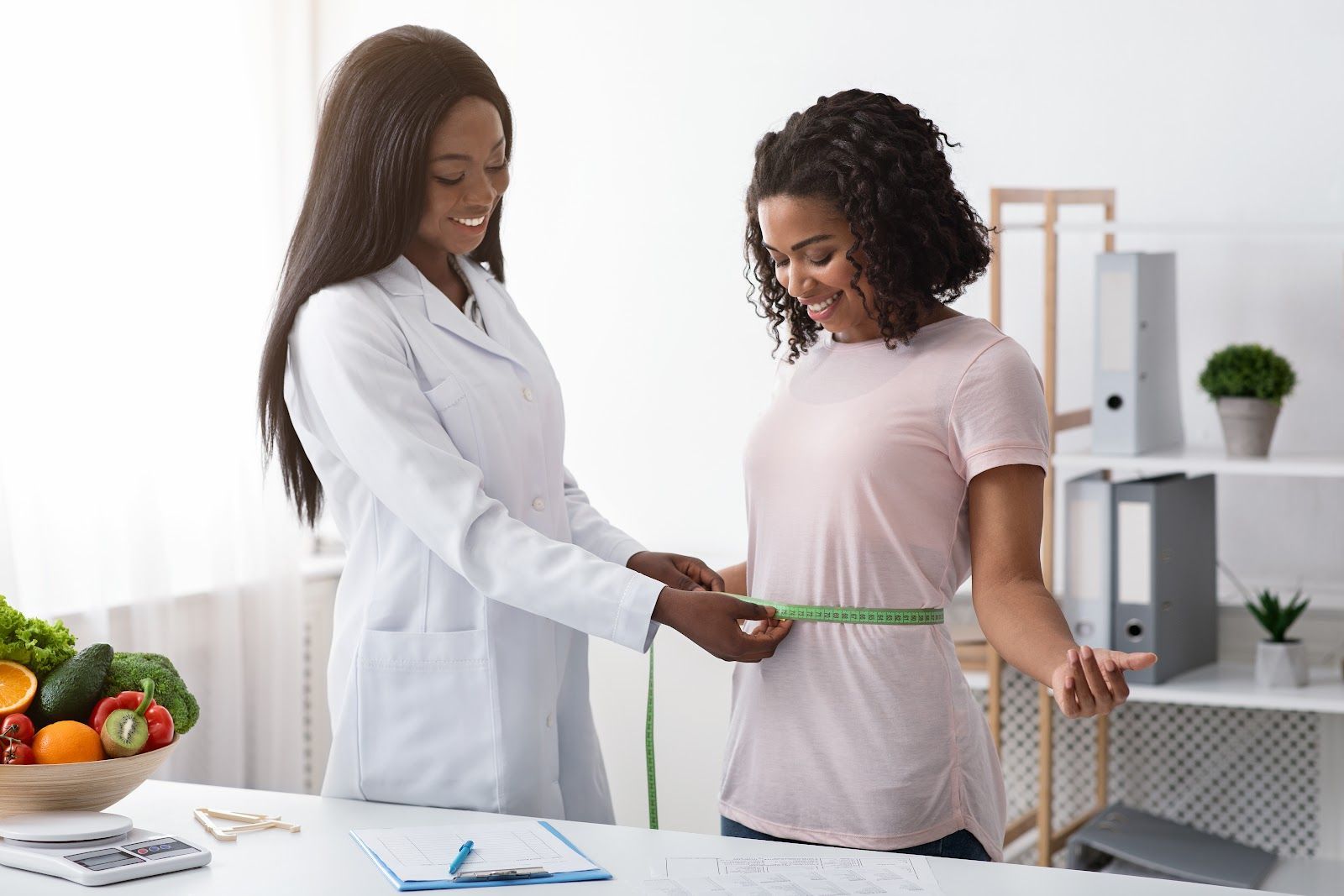 A healthcare provider measures a patient's waist with a measuring tape in an office setting.