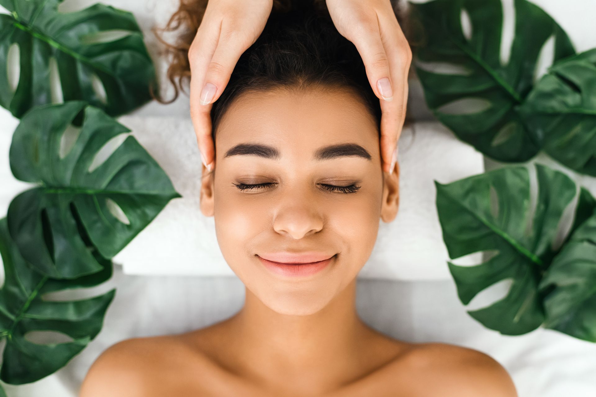 Woman receiving a head massage with closed eyes, surrounded by green leaves.