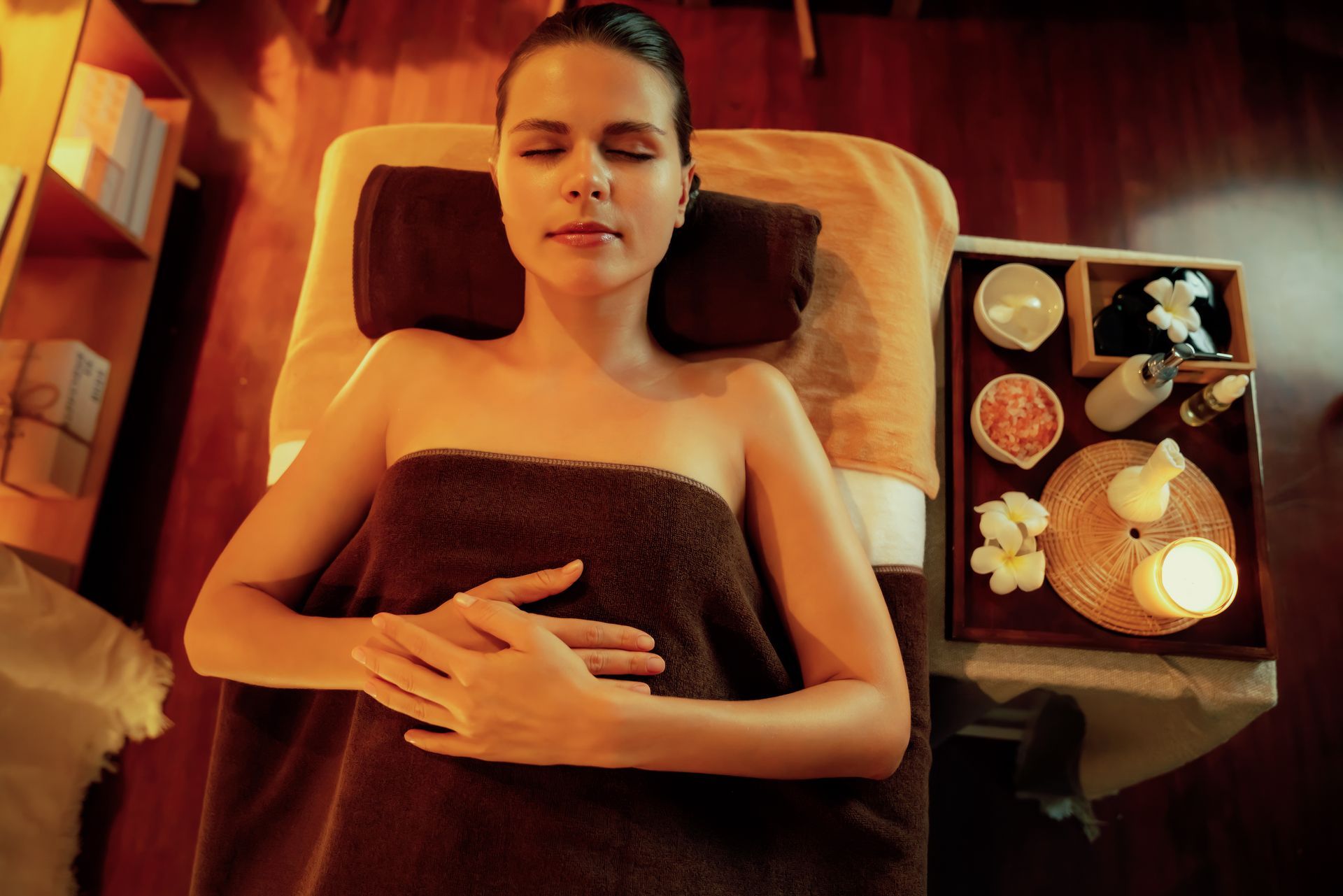 A person lies relaxed on a massage table under a brown towel, next to a tray with spa supplies, candles, and flowers.